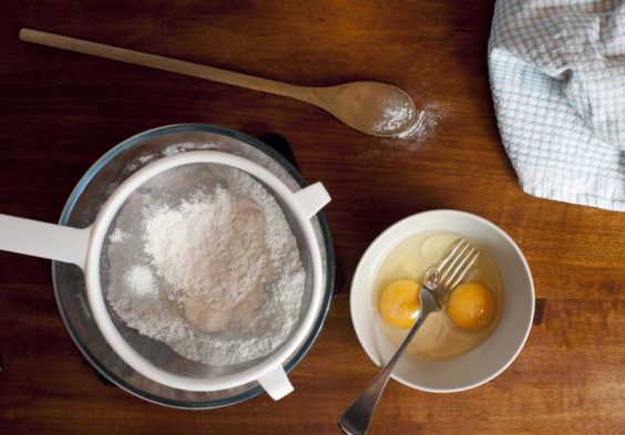 Overhead view of baking ingredients with cake flour in a sieve ready for sifting and two broken eggs, all on a wooden surface